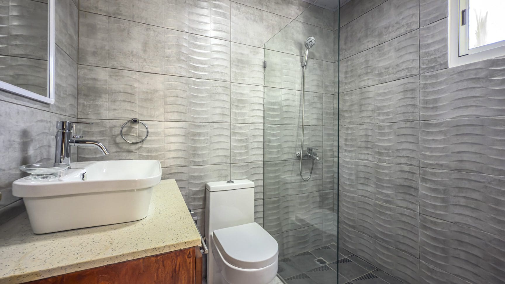 buyDRproperty Modern bathroom with textured gray tiles, white toilet, sink, and glass shower enclosure. Sosua Real Estate