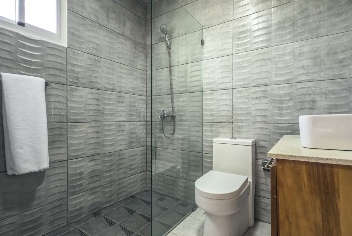 buyDRproperty Modern bathroom with textured gray tile, glass shower, toilet, and white sink on wood cabinet. Sosua Real Estate