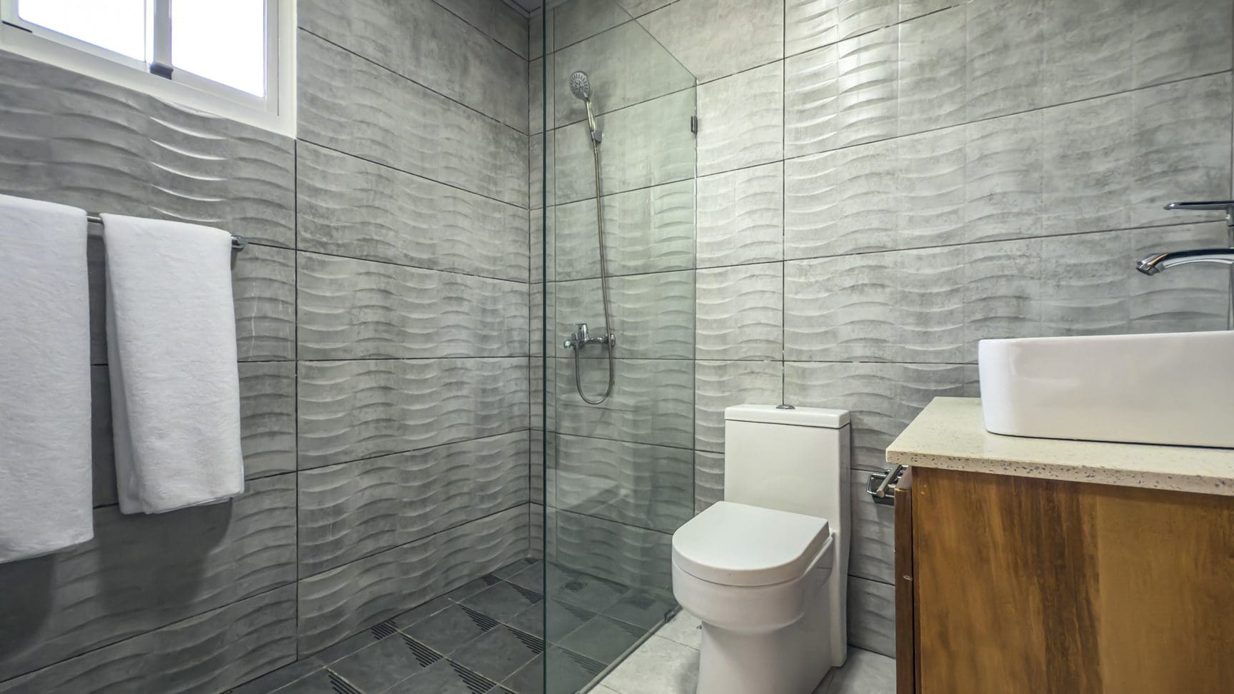buyDRproperty Modern bathroom with textured gray tile, glass shower, toilet, and white sink on wood cabinet. Sosua Real Estate