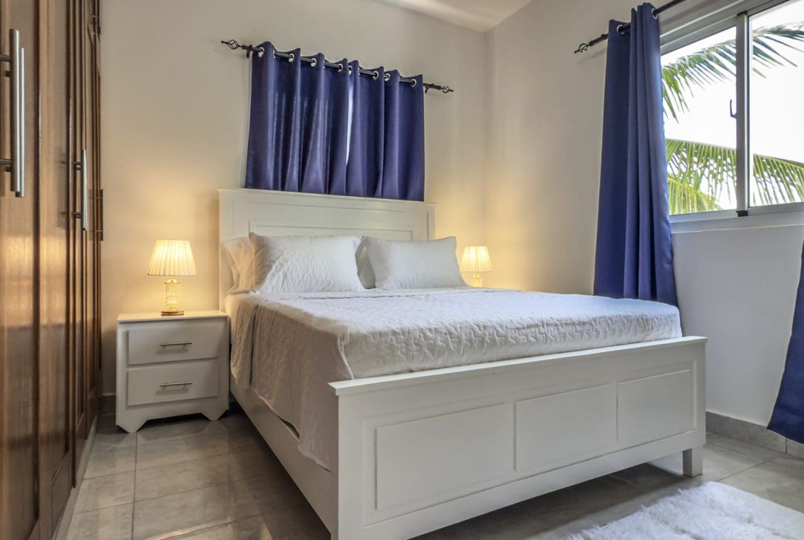 buyDRproperty Bright bedroom with white bed, blue curtains, and wood closet. Sosua Real Estate