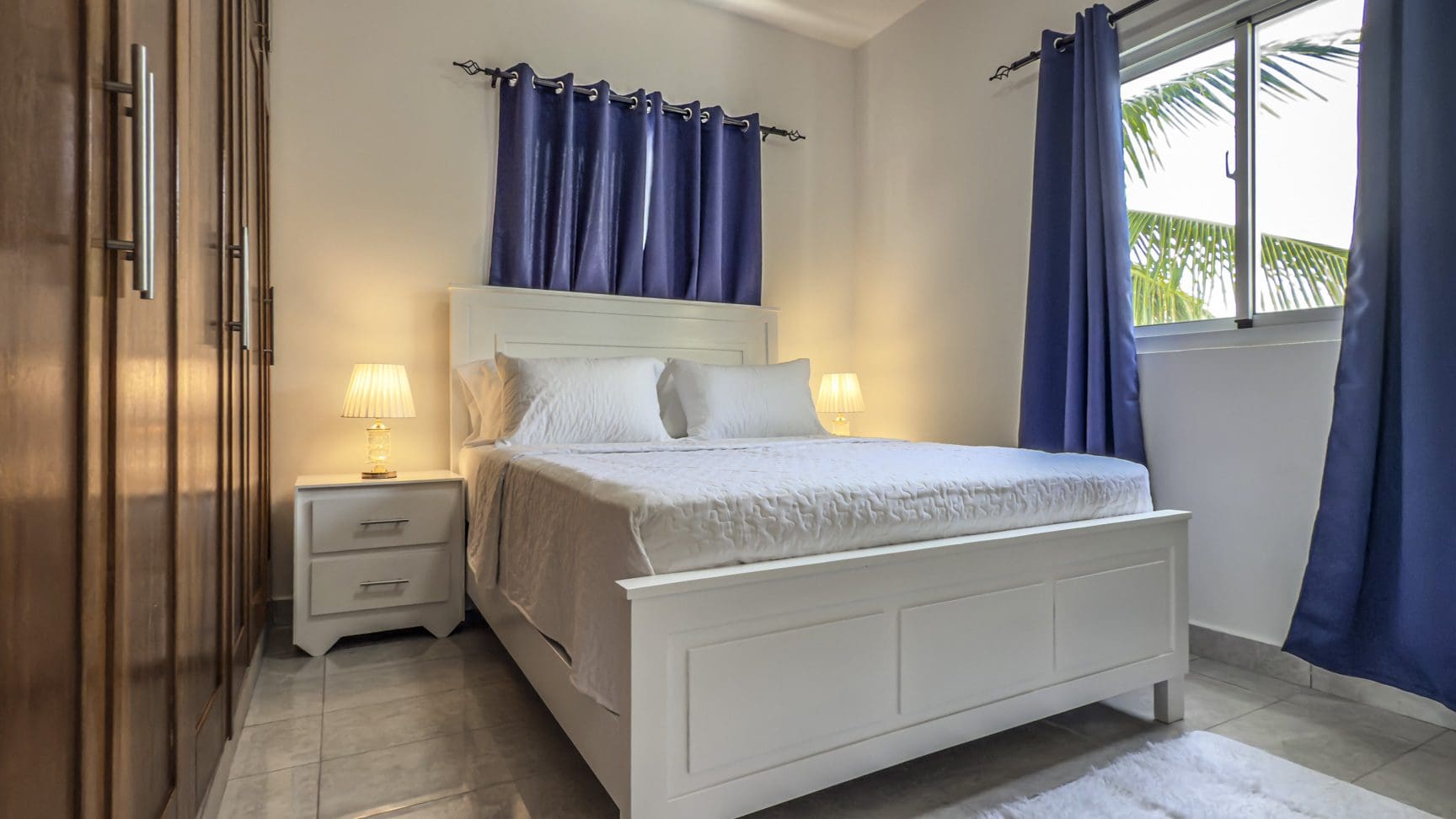 buyDRproperty Bright bedroom with white bed, blue curtains, and wood closet. Sosua Real Estate