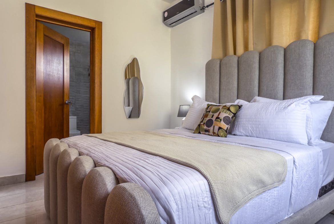 buyDRproperty Modern bedroom with a tufted headboard and footboard, open bathroom door, and minimalist decor. Sosua Real Estate