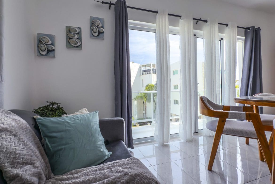 buyDRproperty Bright living room with gray sofa, teal pillow, and dining area by window with white and gray curtains. Sosua Real Estate
