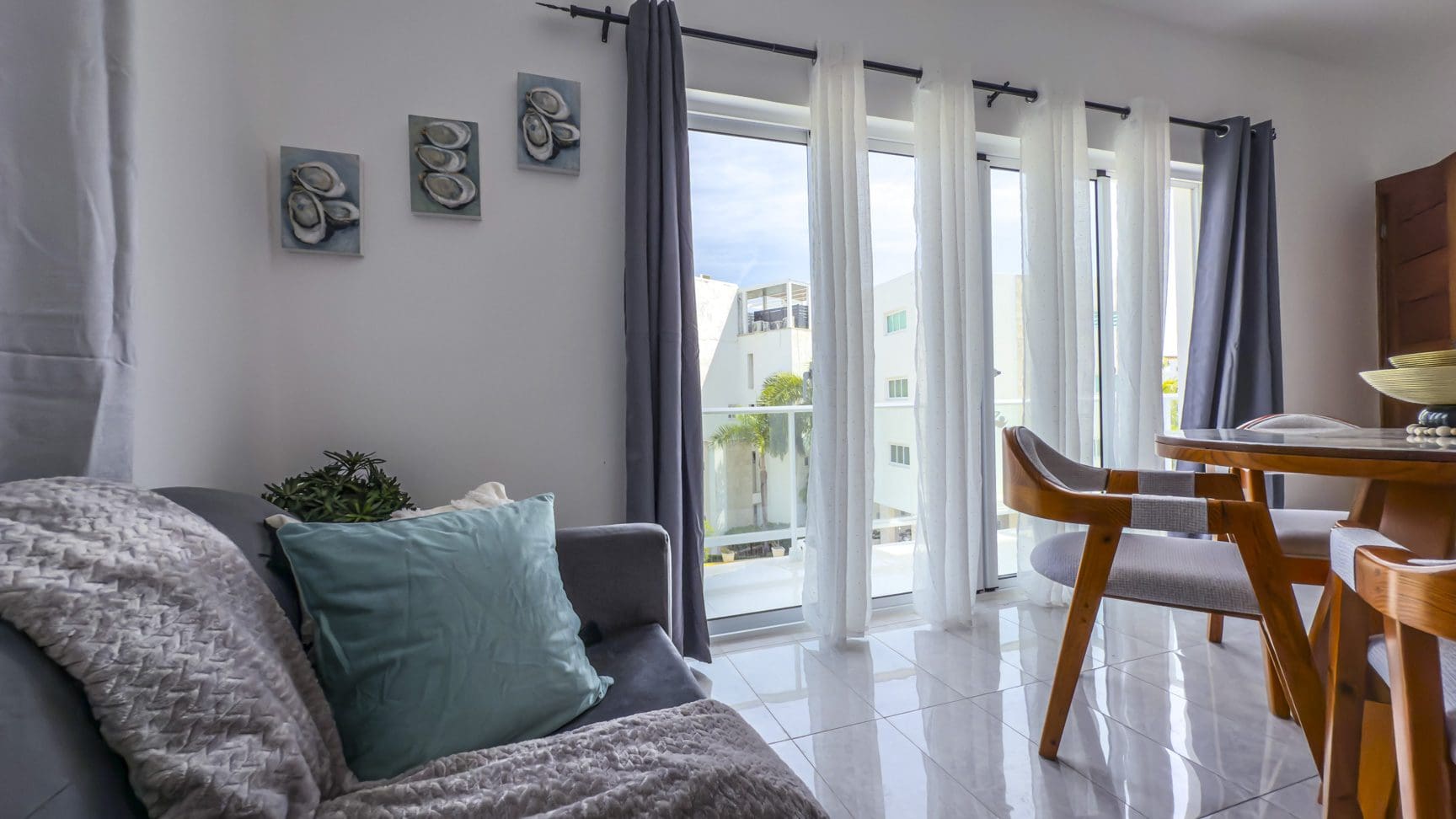 buyDRproperty Bright living room with gray sofa, teal pillow, and dining area by window with white and gray curtains. Sosua Real Estate