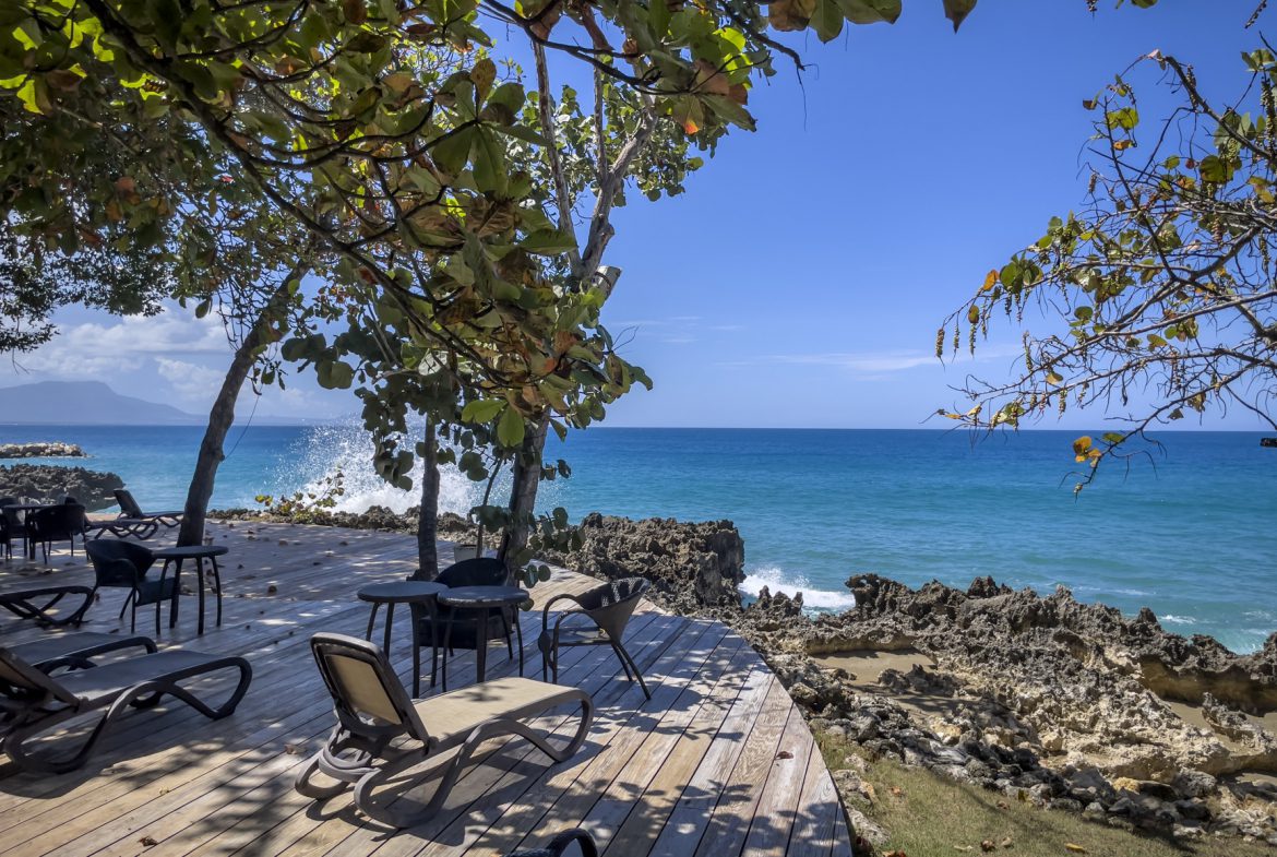buyDRproperty Wooden deck with lounge chairs overlooking a turquoise ocean and rocky coastline under a clear blue sky. Sosua Real Estate