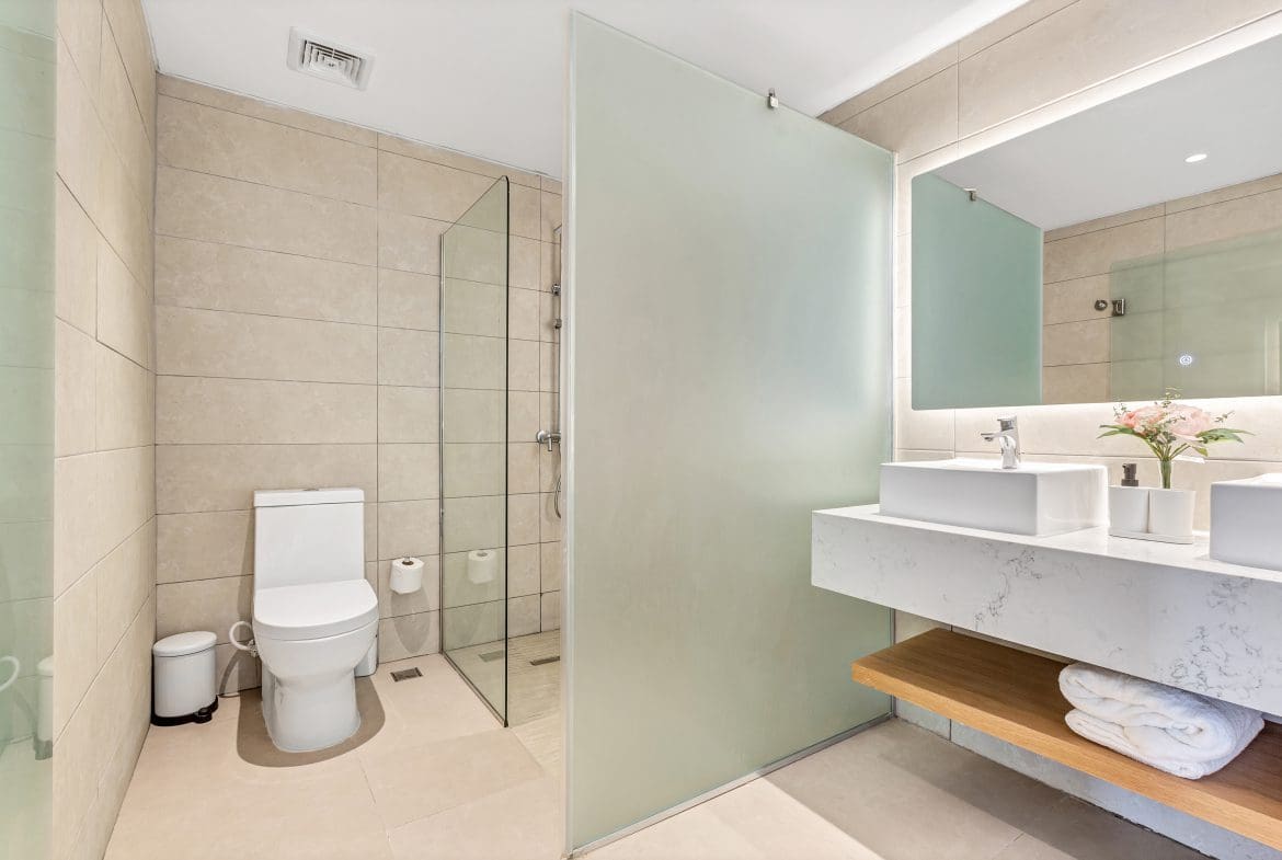 buyDRproperty Modern bathroom with beige tile, white toilet, glass shower, and floating vanity with double sinks. Sosua Real Estate