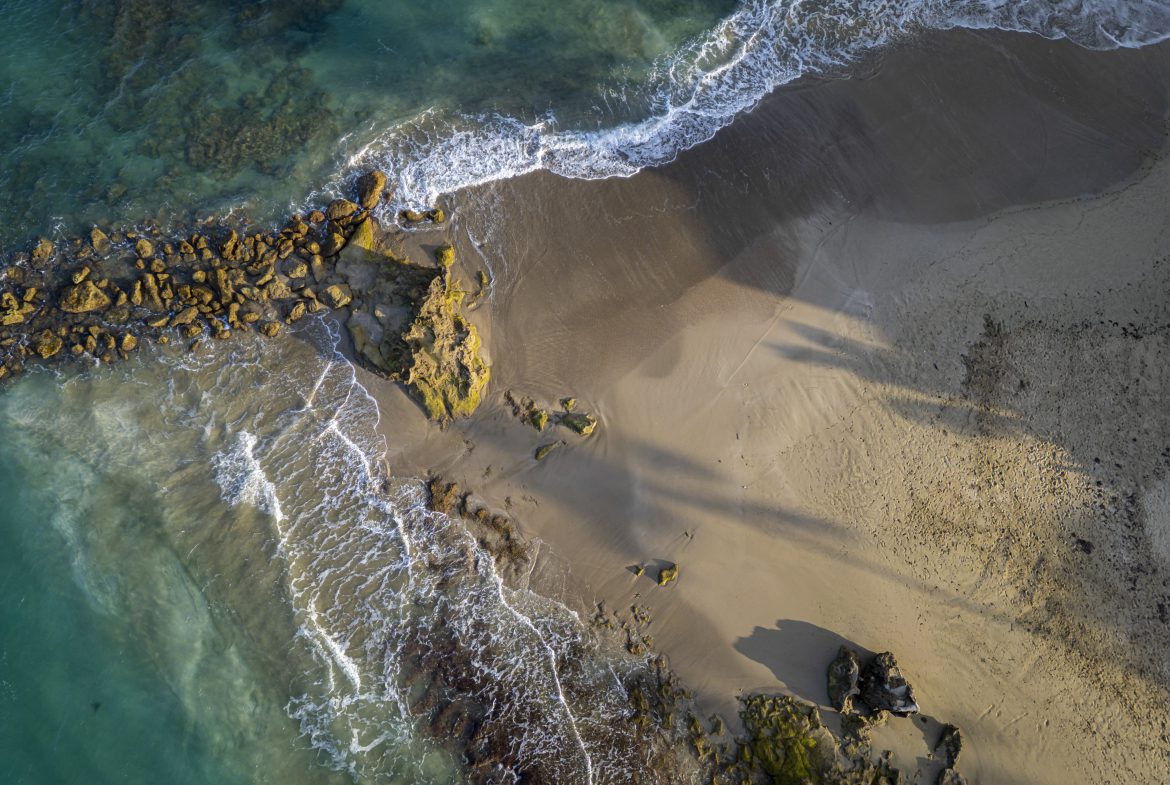 buyDRproperty Aerial view of a tropical beach with turquoise water, rocks, and sandy shore. Sosua Real Estate