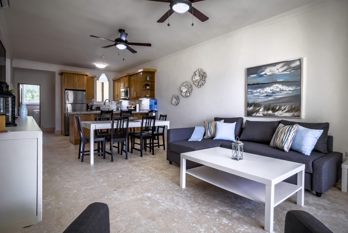 buyDRproperty Living room with gray sofa, white coffee table, dining area, and kitchen. Ceiling fans provide air circulation. Sosua Real Estate