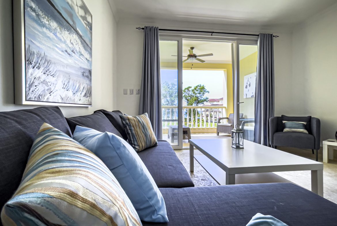 buyDRproperty Cozy living room with gray sofa, blue pillows, and balcony view of tropical trees. Sosua Real Estate