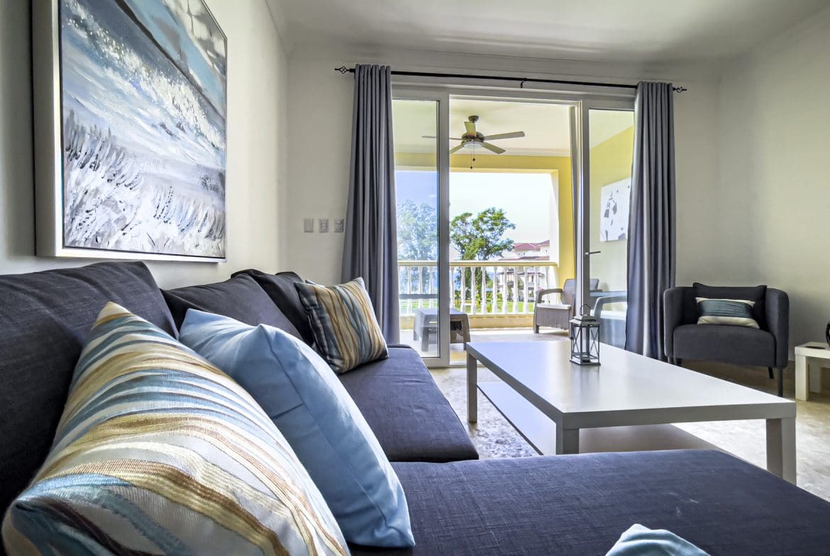 buyDRproperty Cozy living room with gray sofa, blue pillows, and balcony view of tropical trees. Sosua Real Estate