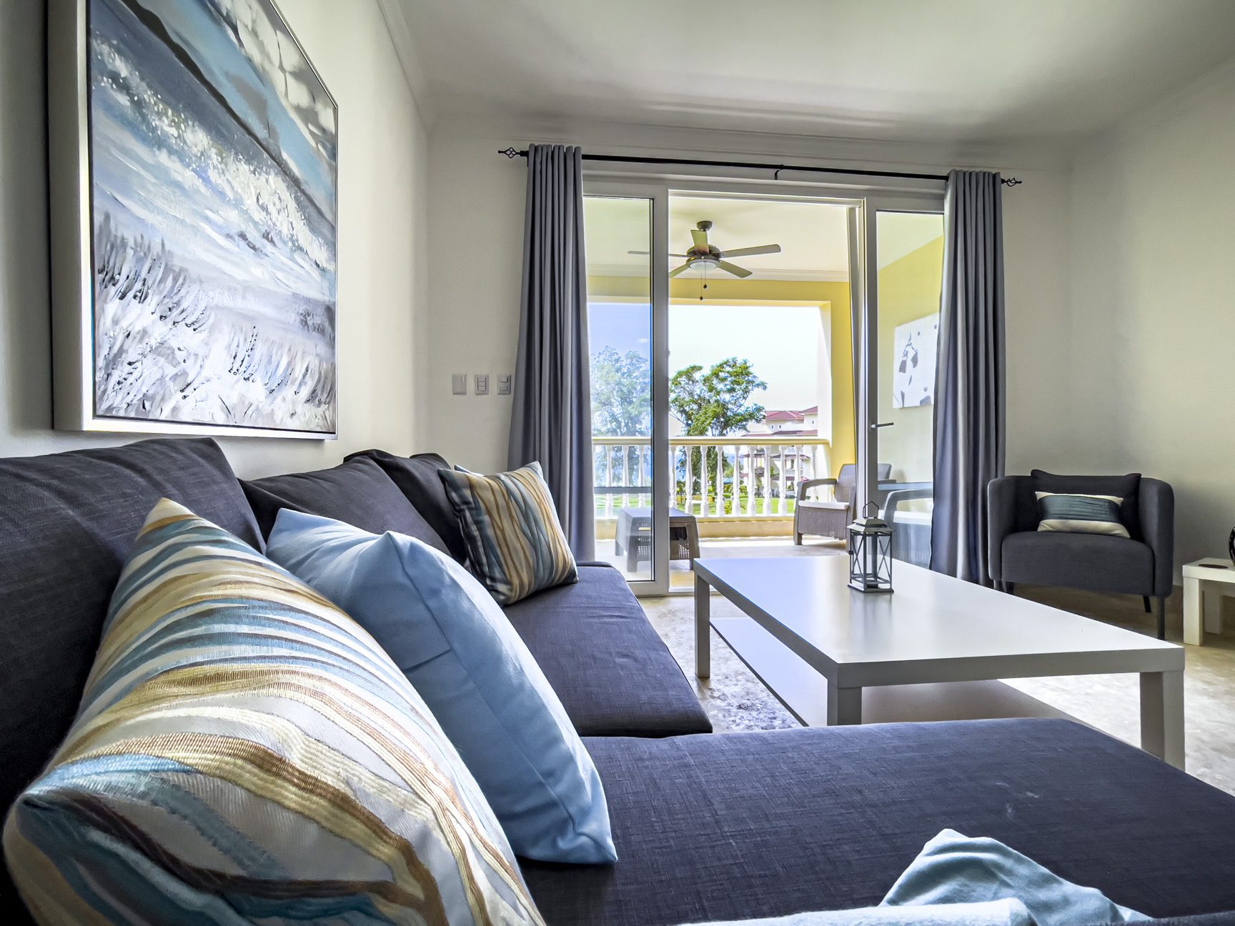 buyDRproperty Cozy living room with gray sofa, blue pillows, and balcony view of tropical trees. Sosua Real Estate
