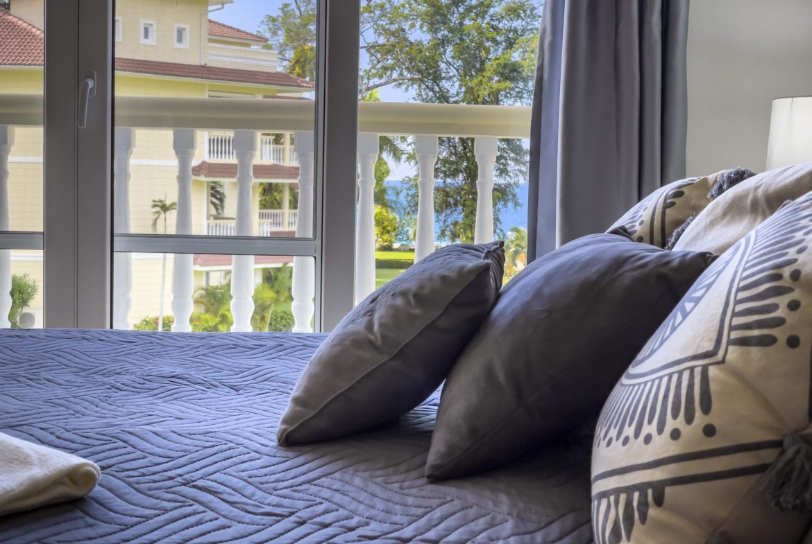 buyDRproperty Bright bedroom with a view of a tropical resort, featuring stylish pillows and a cozy blue quilt. Sosua Real Estate