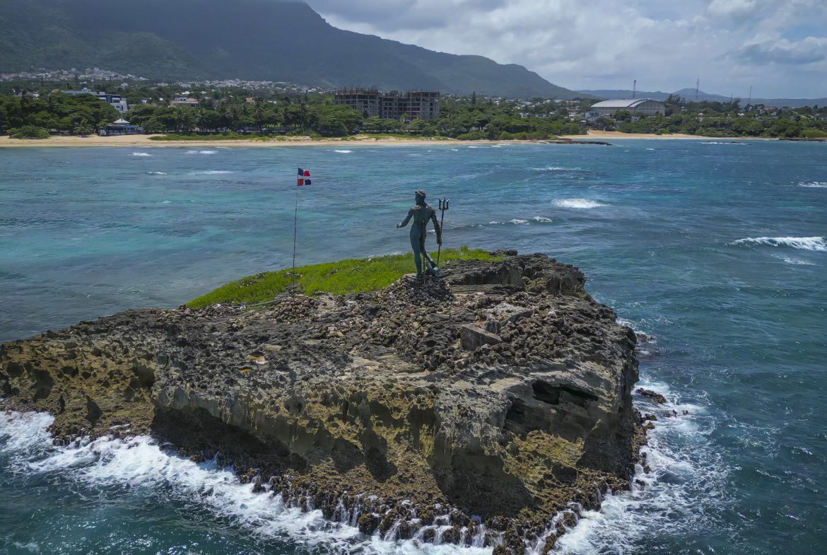 buyDRproperty Statue of Neptune on a rocky island in Sosúa, Dominican Republic, with a flag and the coastline in the background. Sosua Real Estate