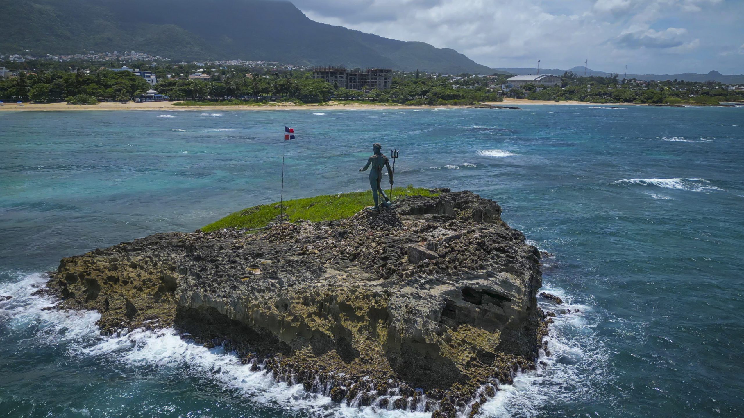 buyDRproperty Statue of Neptune on a rocky island in Sosúa, Dominican Republic, with a flag and the coastline in the background. Sosua Real Estate