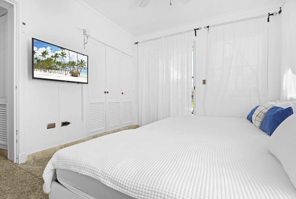 buyDRproperty Bright bedroom with white bedding, TV showing a tropical beach scene, and sheer curtains. Sosua Real Estate