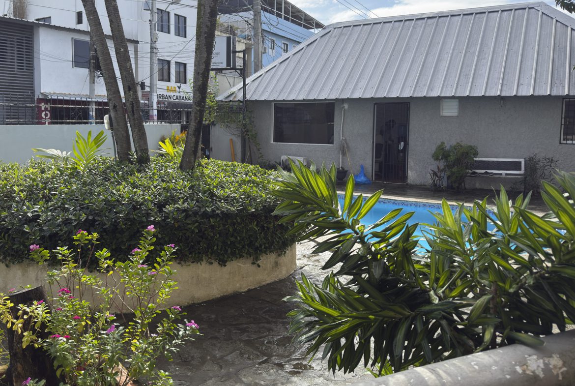 buyDRproperty Lush tropical garden with pool, gray building, and trees under a bright sky. Sosua Real Estate