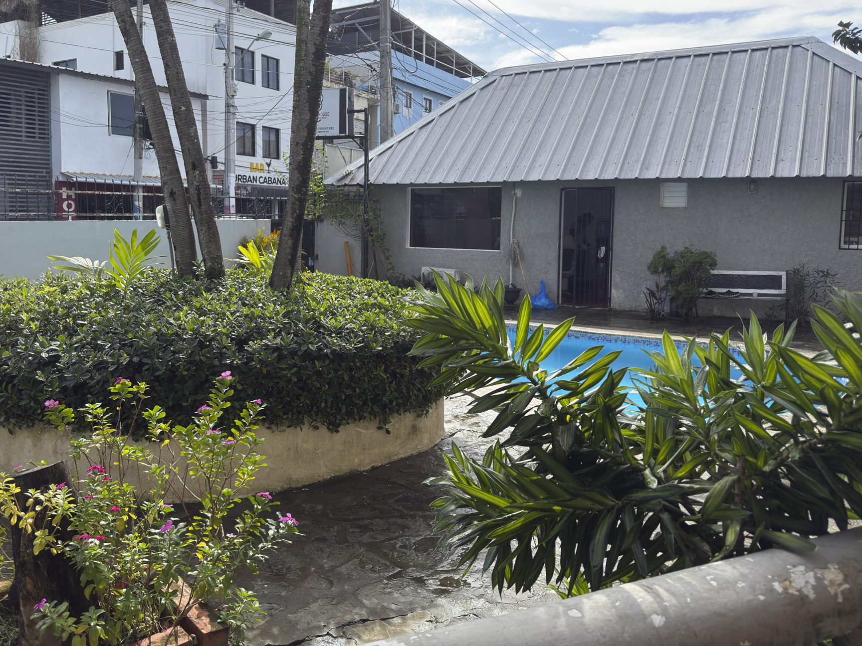 buyDRproperty Lush tropical garden with pool, gray building, and trees under a bright sky. Sosua Real Estate