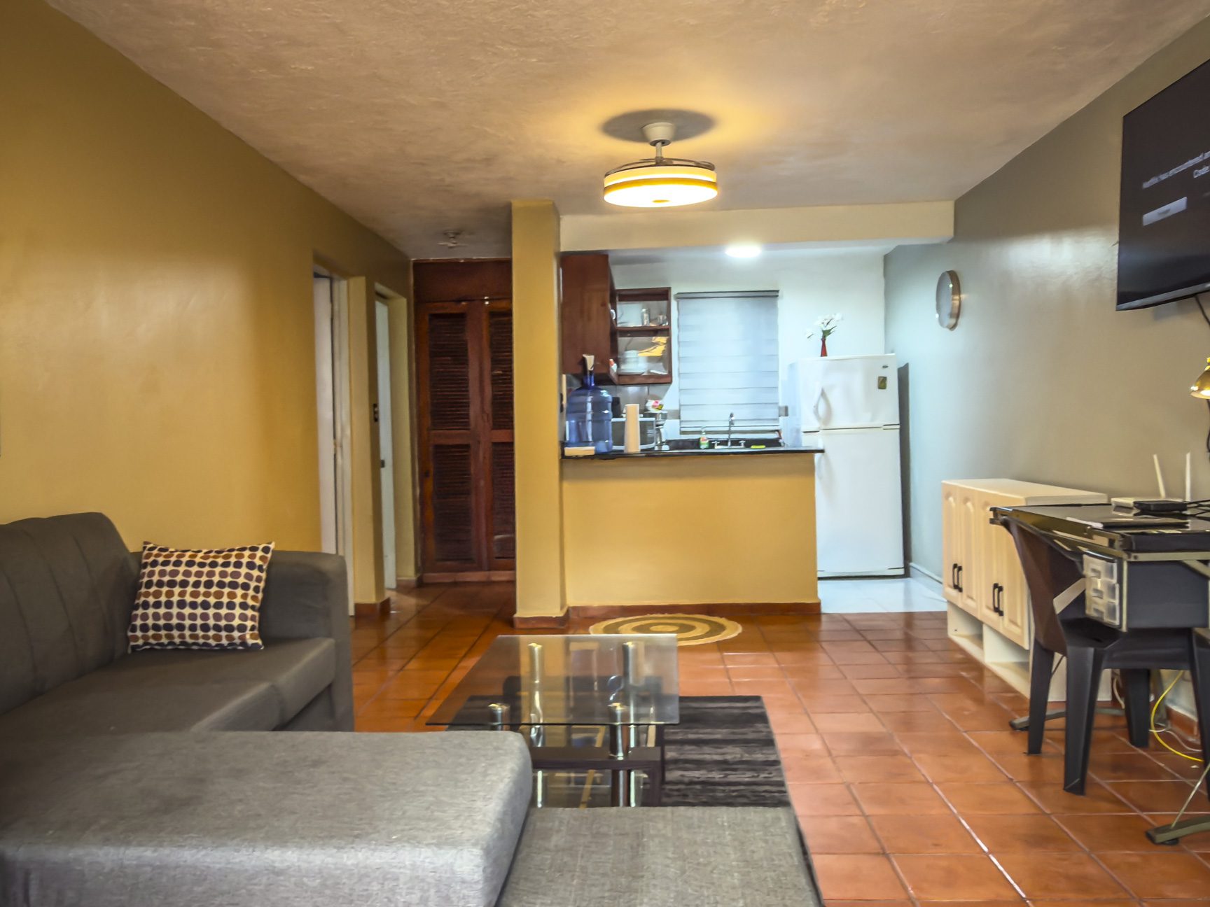 buyDRproperty Cozy apartment interior with a gray sectional sofa, glass coffee table, and a compact kitchen area. Sosua Real Estate