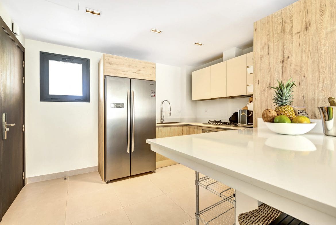 buyDRproperty Modern kitchen with stainless steel fridge, white countertops, light wood cabinets, and fresh tropical fruit display. Sosua Real Estate