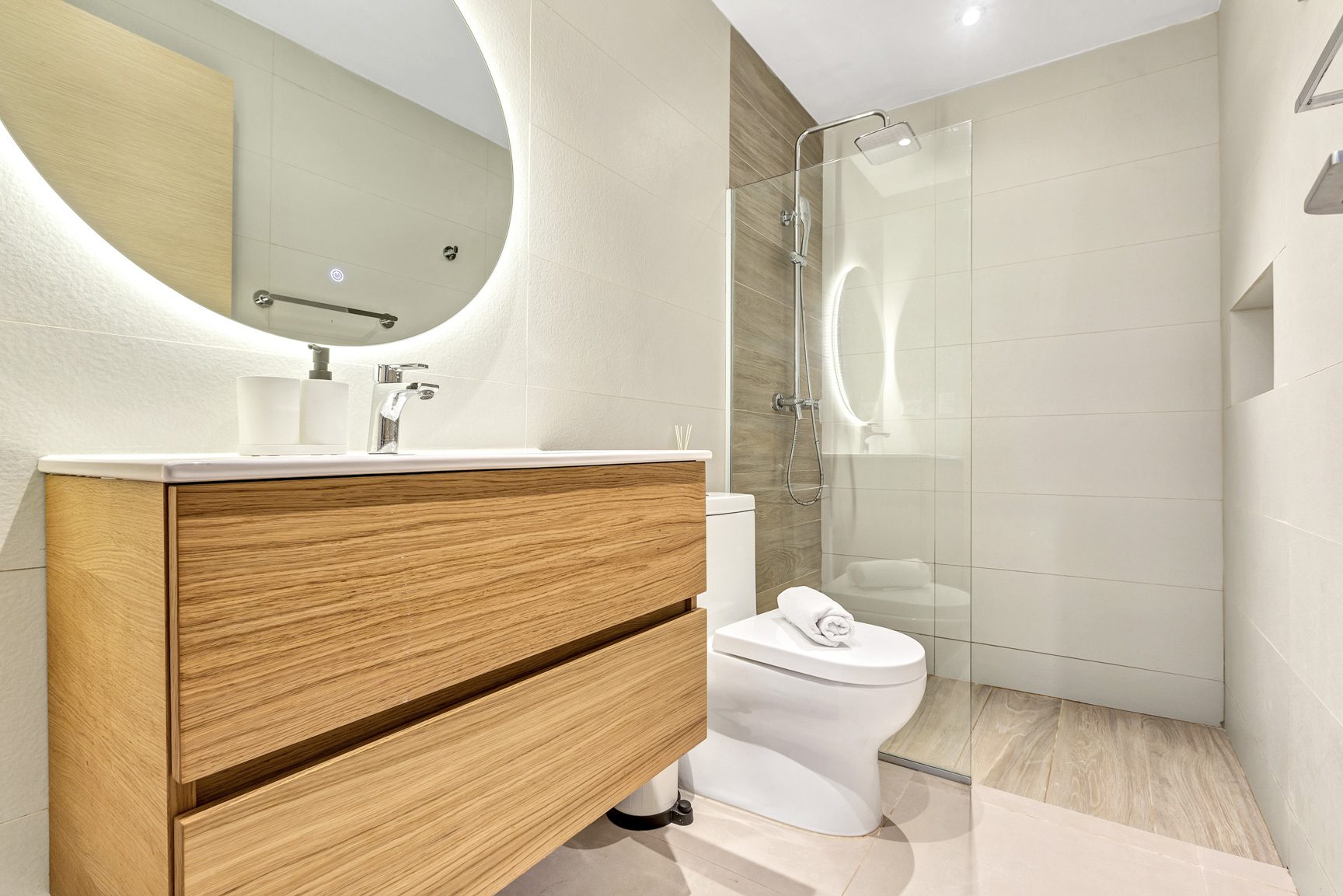 buyDRproperty Modern bathroom with wood vanity, round lighted mirror, glass shower, and white toilet. Sosua Real Estate
