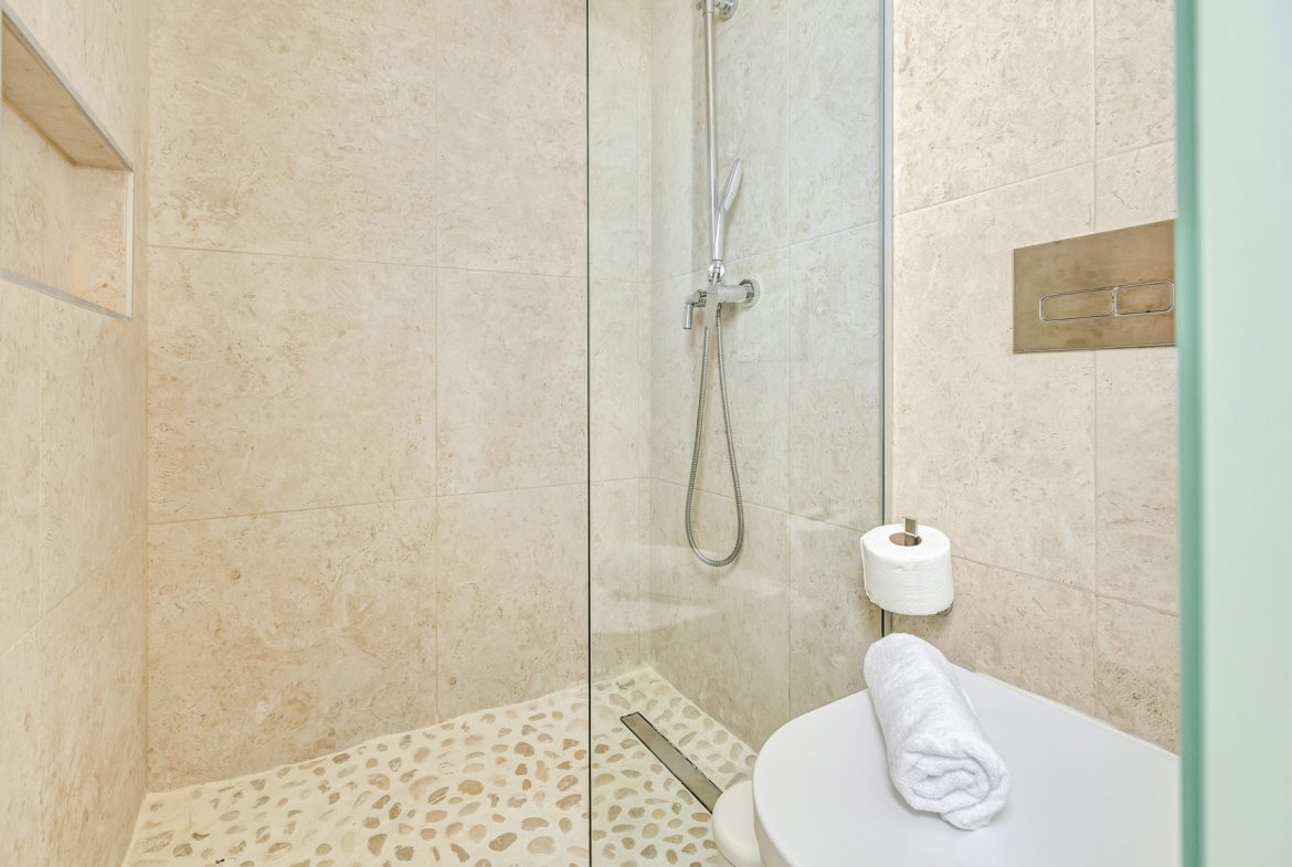 buyDRproperty Modern bathroom with beige tile, glass shower, toilet, and pebble floor. Sosua Real Estate