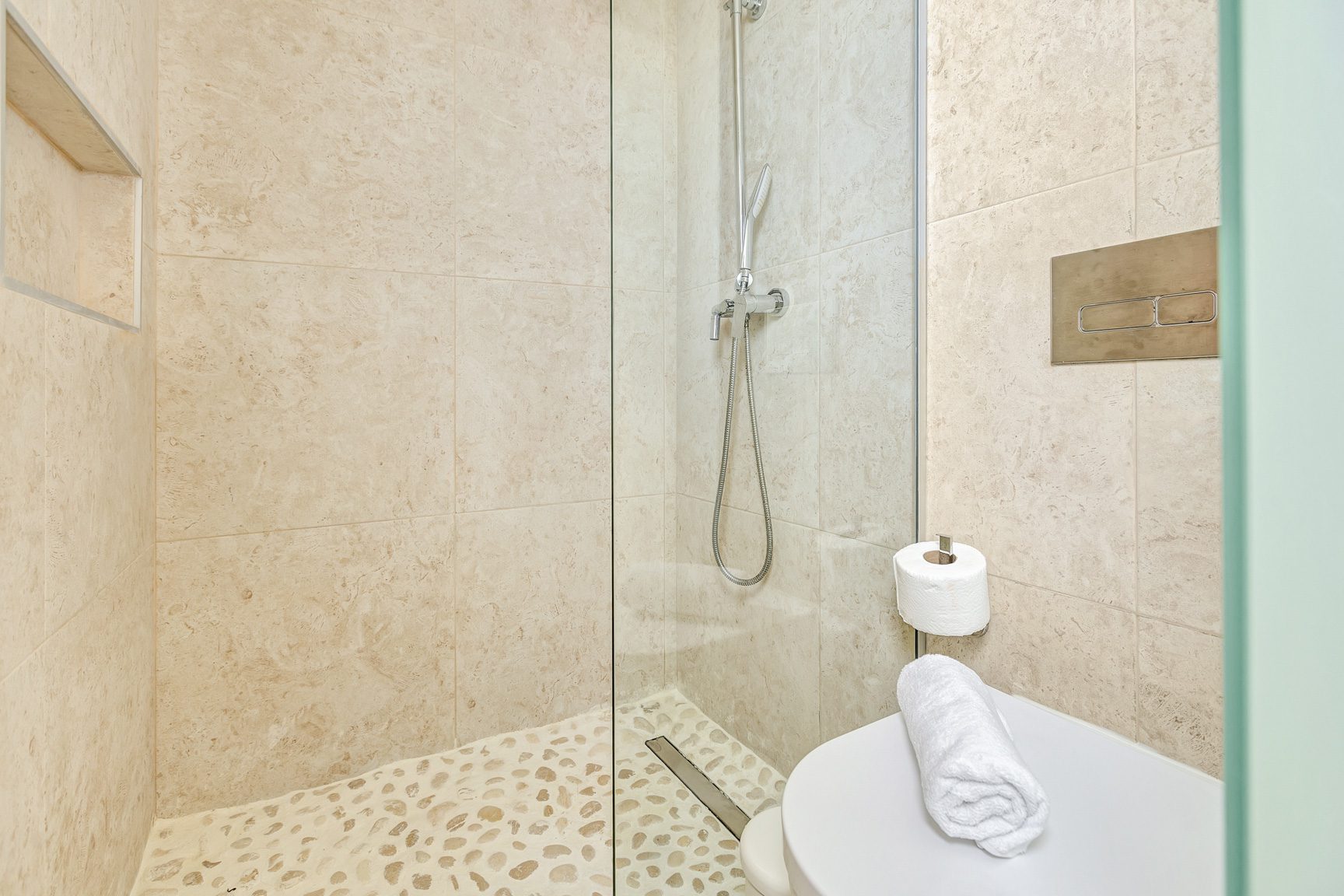 buyDRproperty Modern bathroom with beige tile, glass shower, toilet, and pebble floor. Sosua Real Estate