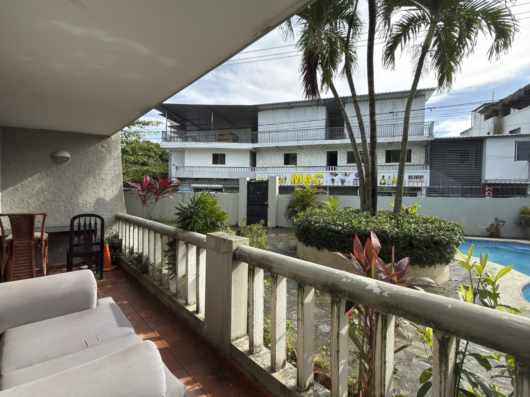 buyDRproperty Balcony view of a building with "MAC Delivery" sign, palm trees, and a pool in a tropical location. Sosua Real Estate