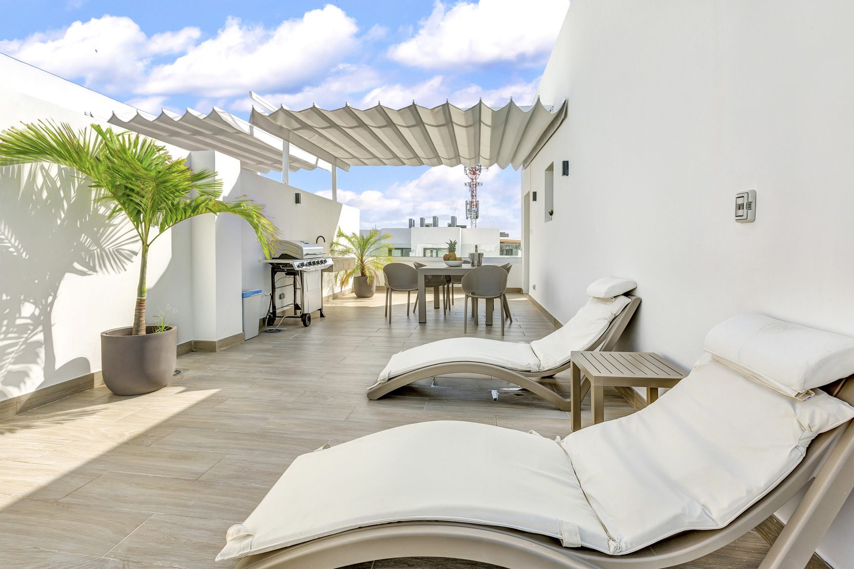 buyDRproperty Luxury rooftop patio with loungers, dining table, grill, and palm tree against a blue sky. Sosua Real Estate
