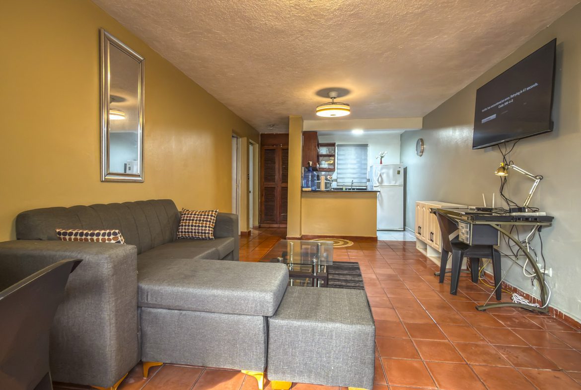 buyDRproperty Cozy living room with gray sectional sofa, terracotta tile floor, and open kitchen in background. Sosua Real Estate