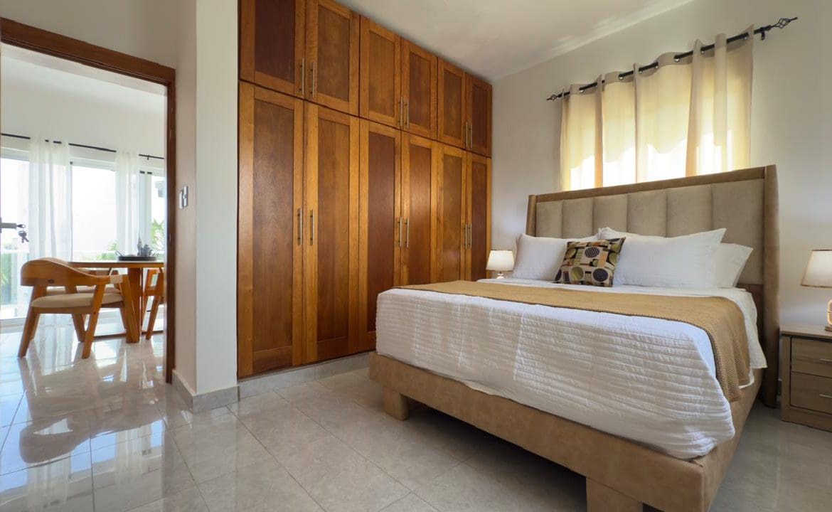 buyDRproperty Bedroom with wooden wardrobe, king-size bed with white linens, and view to a bright dining area. Sosua Real Estate