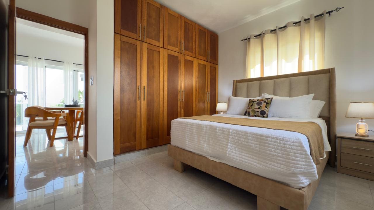 buyDRproperty Bedroom with wooden wardrobe, king-size bed with white linens, and view to a bright dining area. Sosua Real Estate