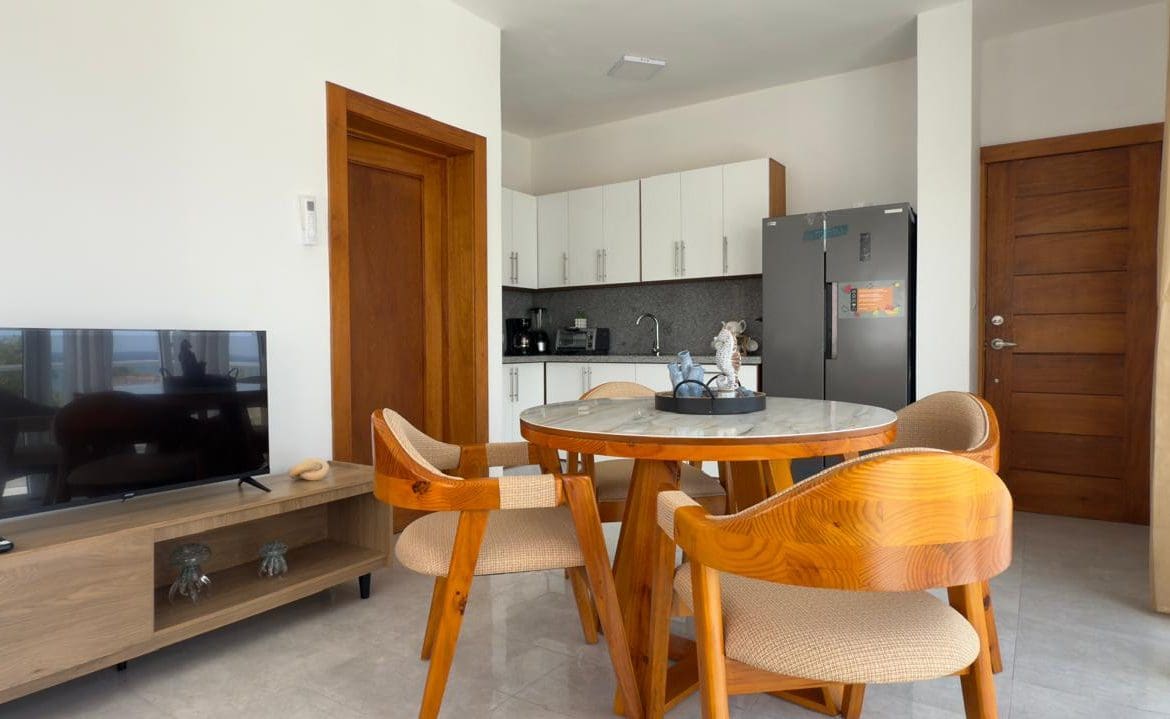 buyDRproperty Modern apartment dining area with round table, chairs, TV, and kitchen. Sosua Real Estate