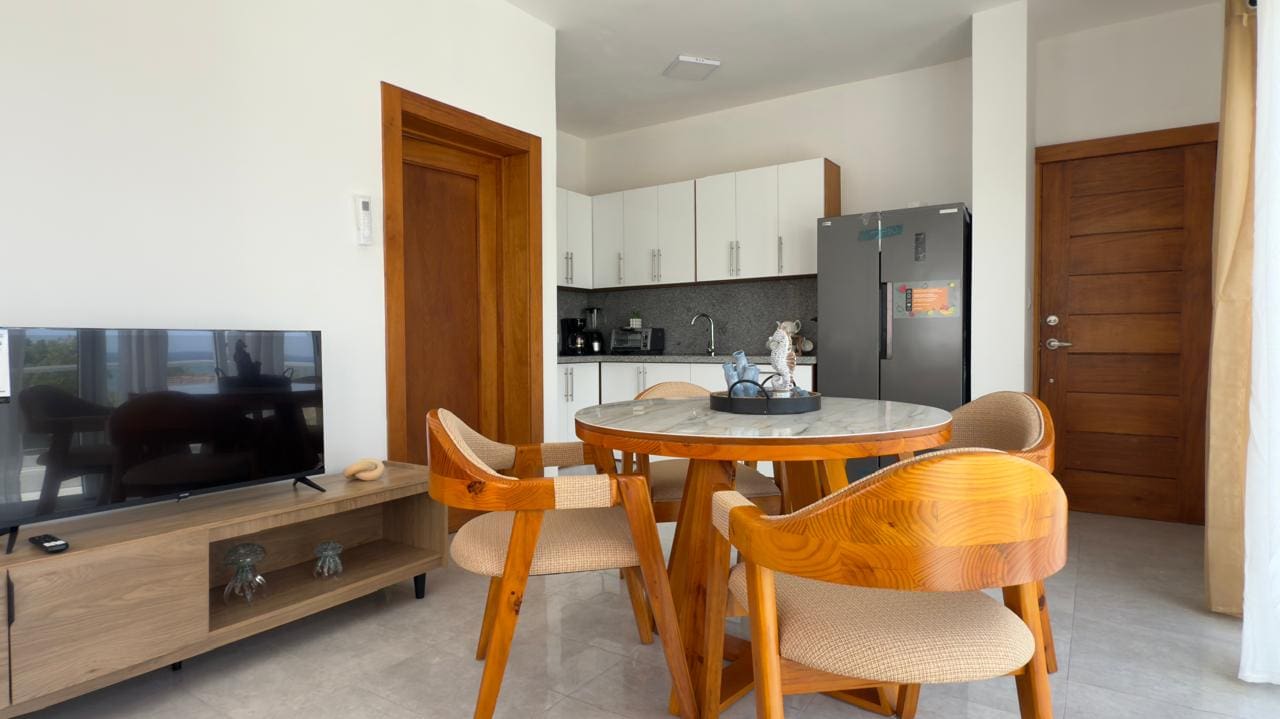 buyDRproperty Modern apartment dining area with round table, chairs, TV, and kitchen. Sosua Real Estate