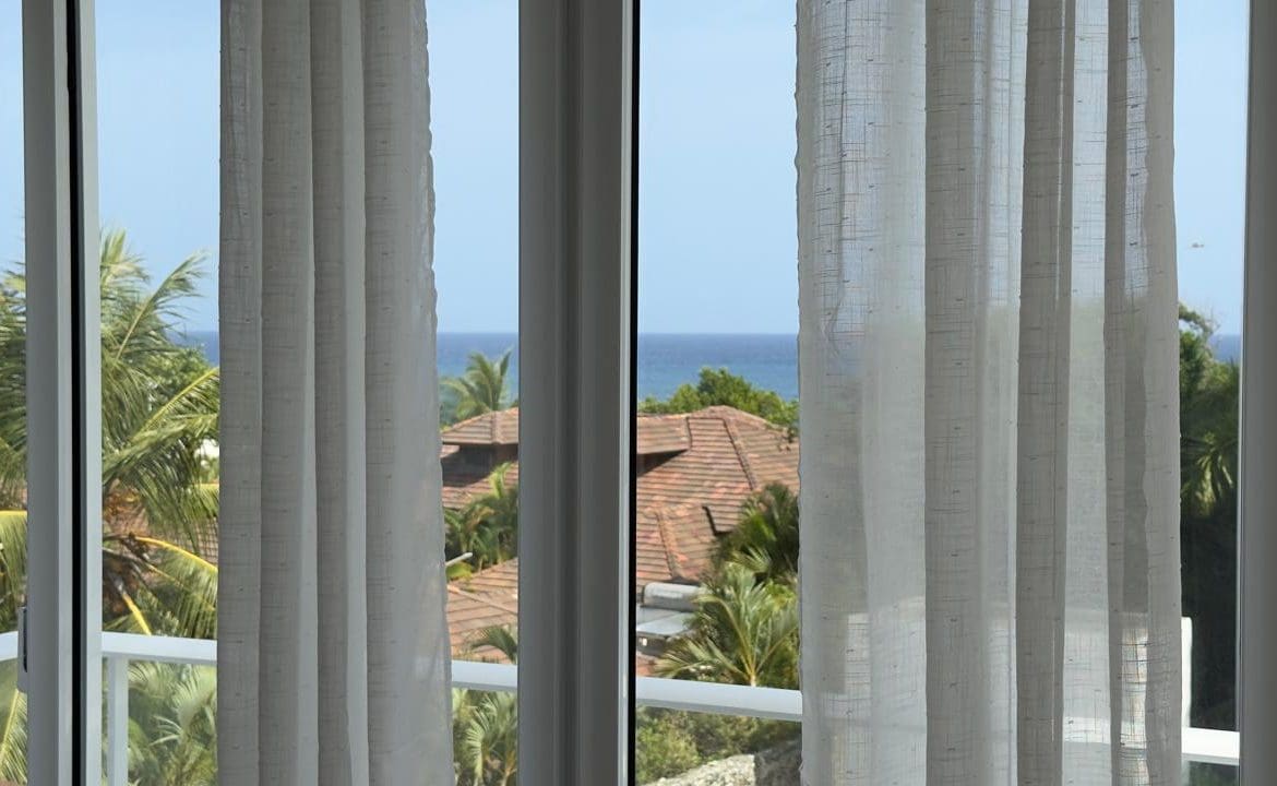 buyDRproperty Ocean view from a window with white curtains, tropical trees, and rooftops. Sosua Real Estate