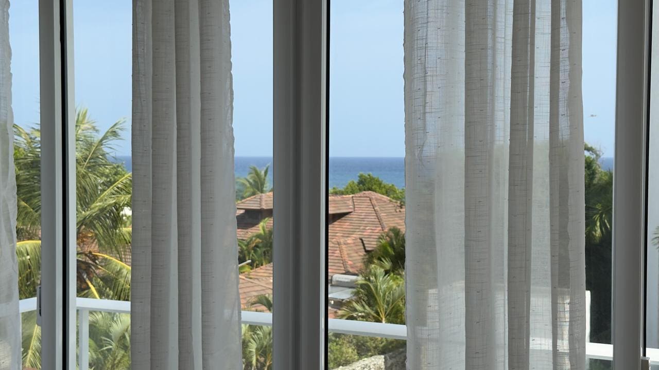 buyDRproperty Ocean view from a window with white curtains, tropical trees, and rooftops. Sosua Real Estate