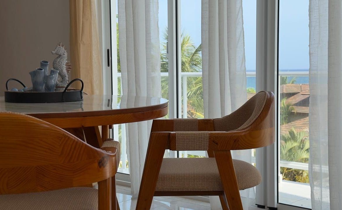 buyDRproperty Dining area with wooden chairs, glass table, and ocean view through sheer curtains. Sosua Real Estate