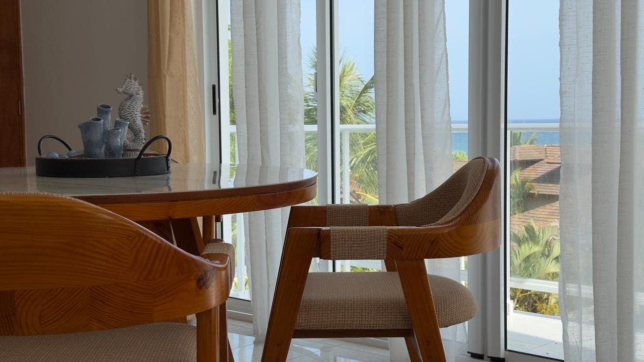 buyDRproperty Dining area with wooden chairs, glass table, and ocean view through sheer curtains. Sosua Real Estate