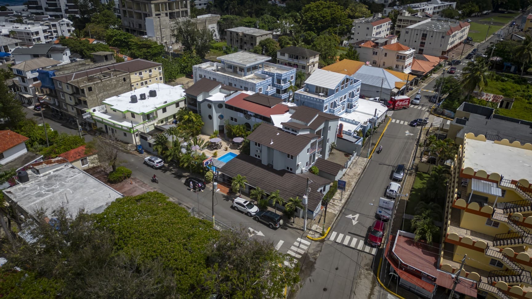 Aerial view of a tropical town street with colorful houses, palm trees, and parked cars.