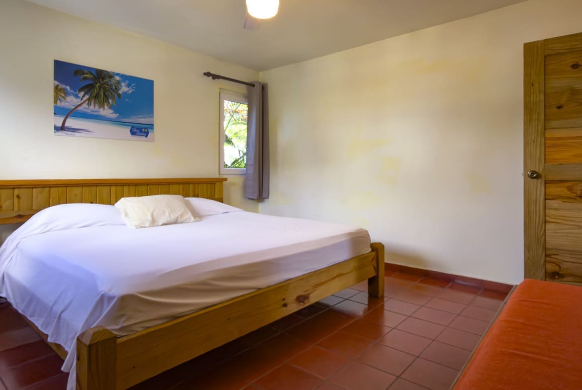 buyDRproperty Cozy bedroom with a wooden bed, white linens, and a tropical palm tree painting. Sosua Real Estate