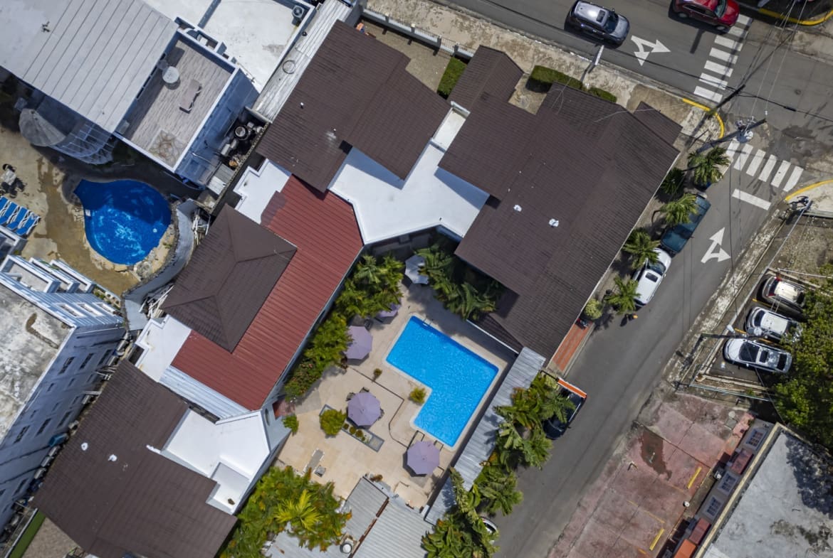 Aerial view of a hotel complex with brown roofs, two blue pools, palm trees, and a surrounding street.