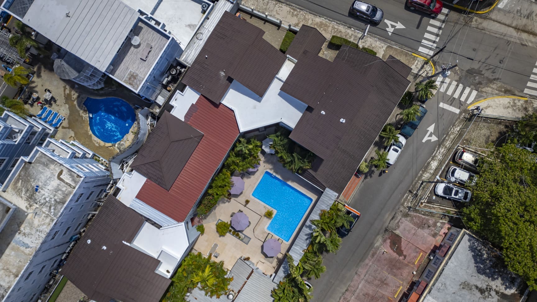 Aerial view of a hotel complex with brown roofs, two blue pools, palm trees, and a surrounding street.