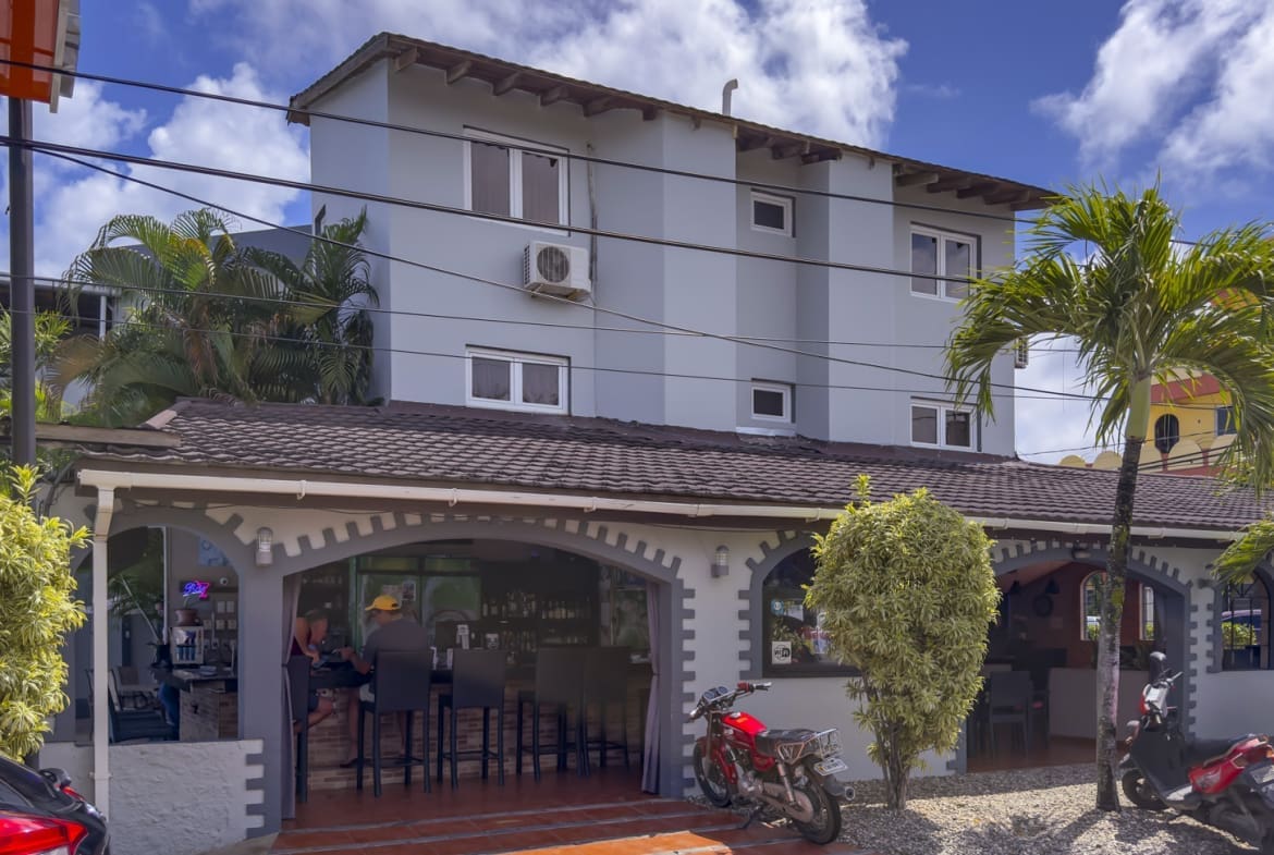 Cafe with outdoor seating under arched porch in front of a light blue building; palm trees, motorcycles, and a parked car outside on a sunny day.