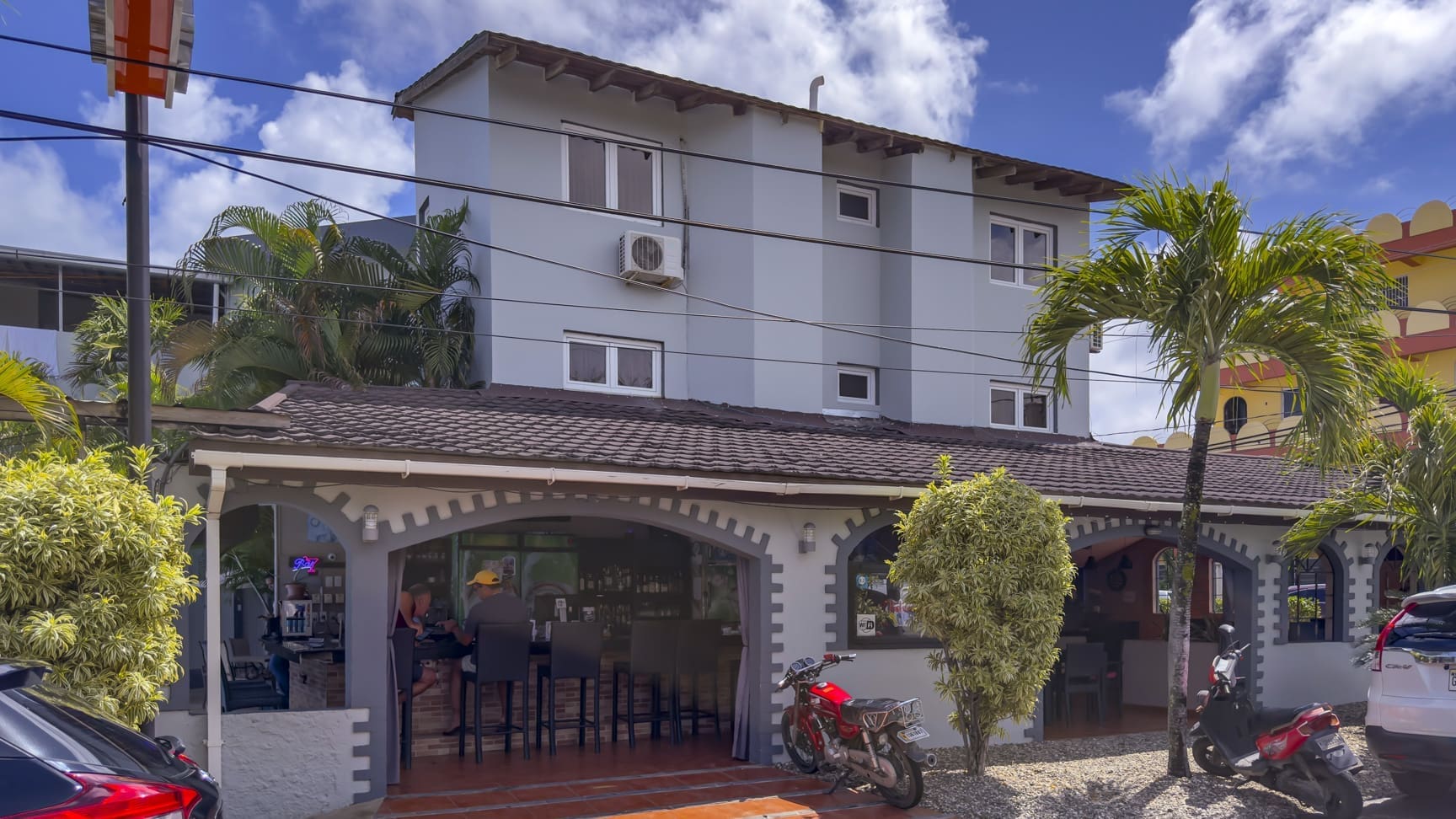 Cafe with outdoor seating under arched porch in front of a light blue building; palm trees, motorcycles, and a parked car outside on a sunny day.