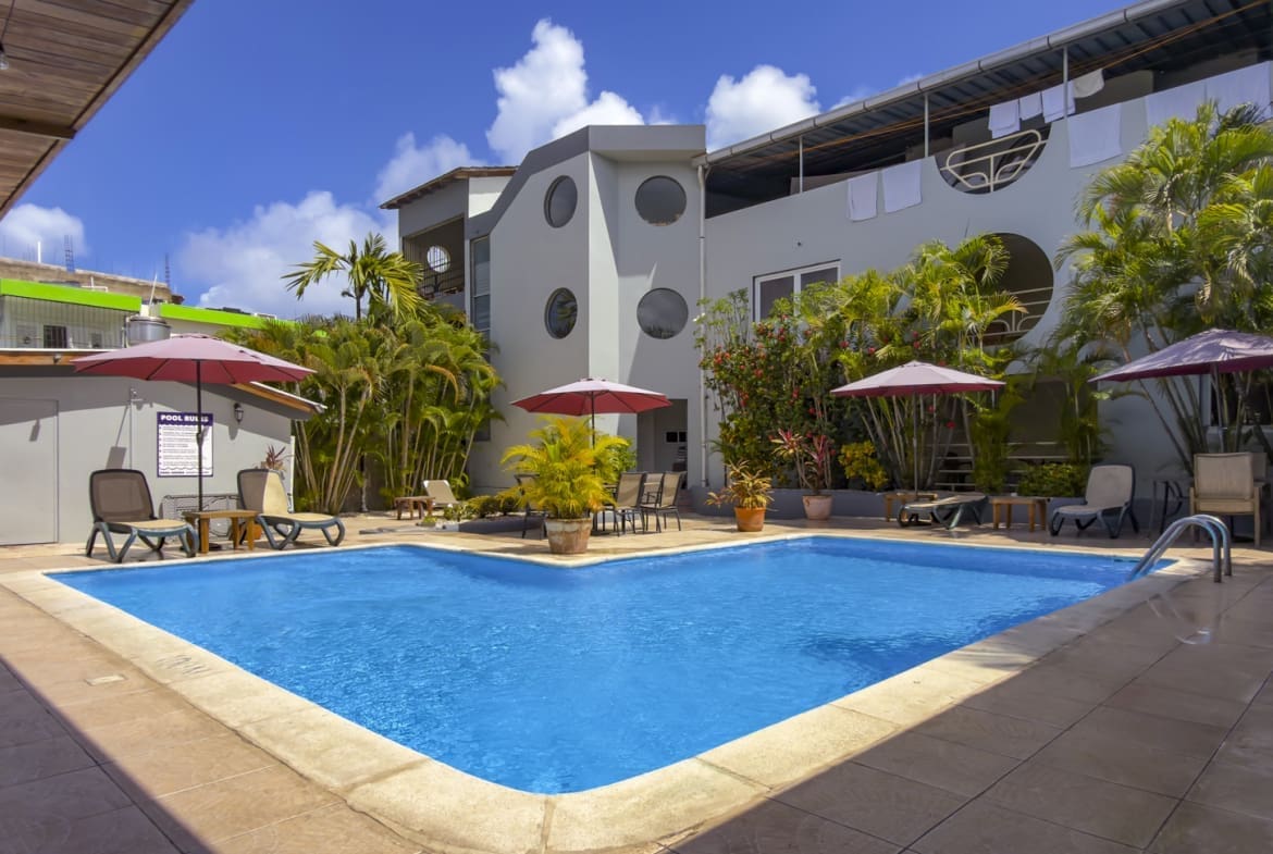 Hotel courtyard pool with blue water, beige deck, pink umbrellas, sun loungers, and tropical plants against a modern white building.