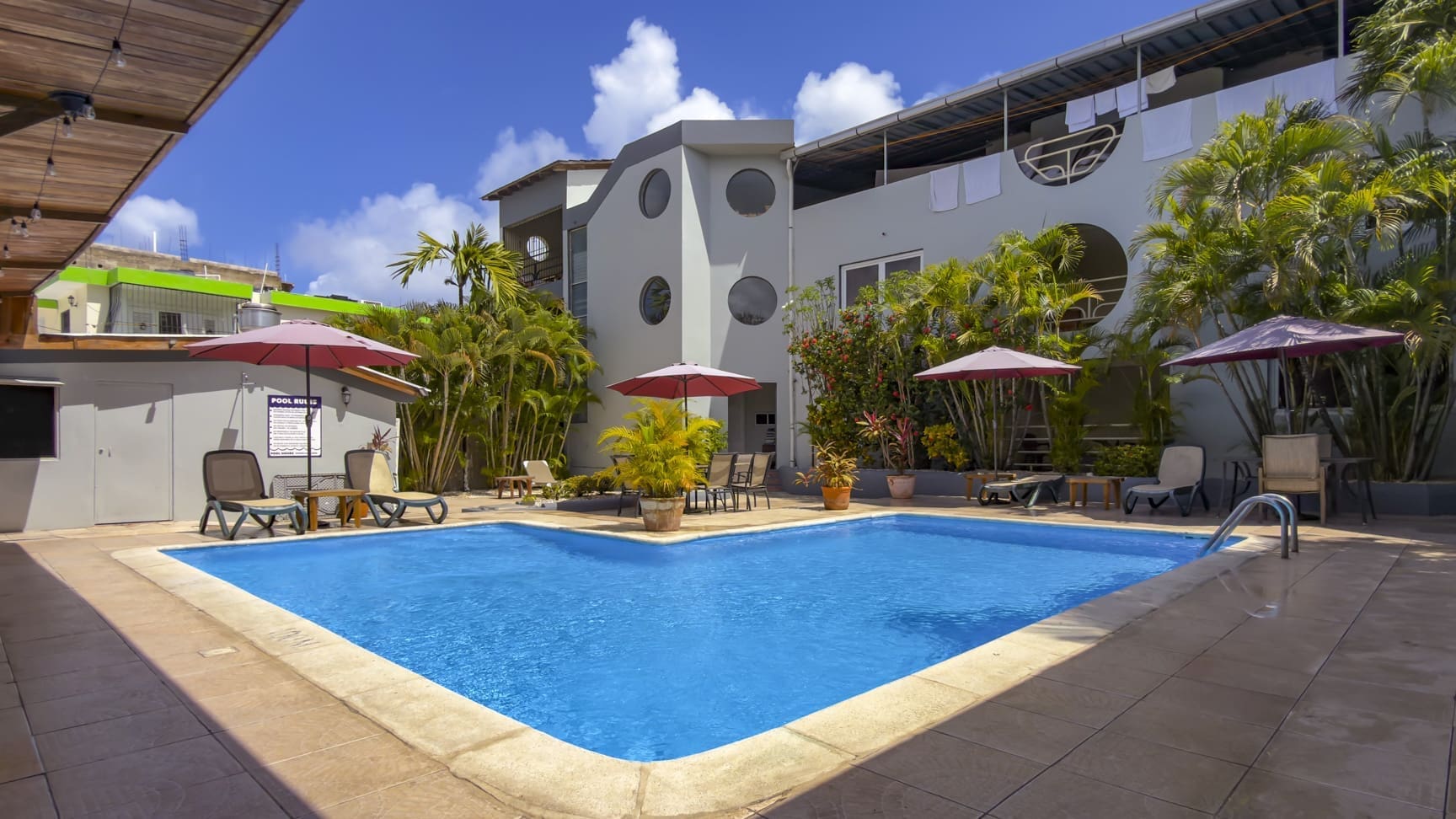 Hotel courtyard pool with blue water, beige deck, pink umbrellas, sun loungers, and tropical plants against a modern white building.