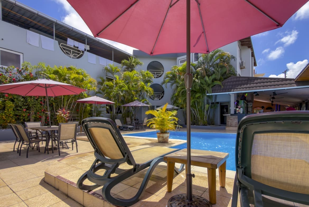 Poolside patio with pink umbrellas, lounge chairs, and tables among tropical plants at a sunny hotel courtyard.