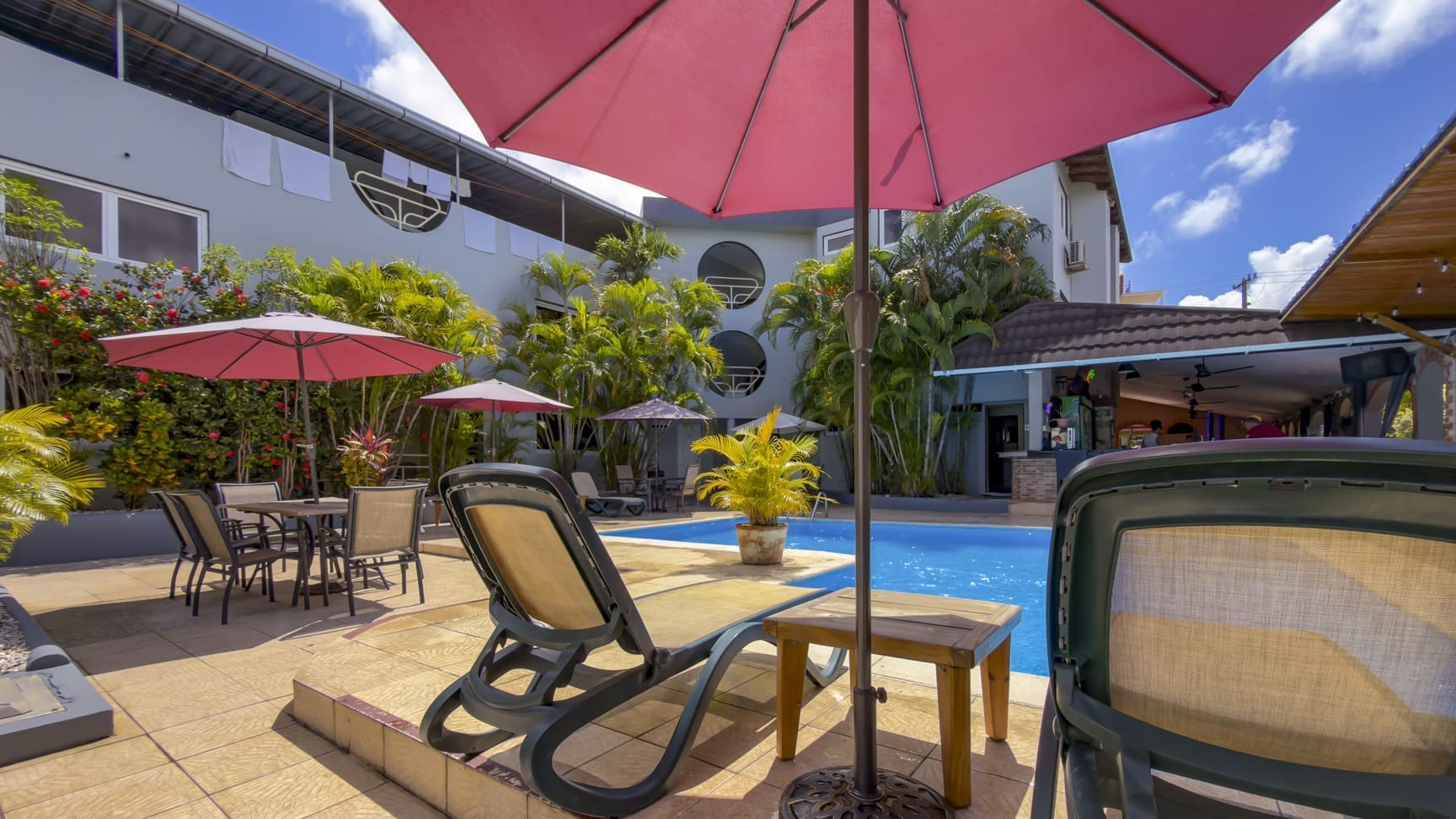 Poolside patio with pink umbrellas, lounge chairs, and tables among tropical plants at a sunny hotel courtyard.