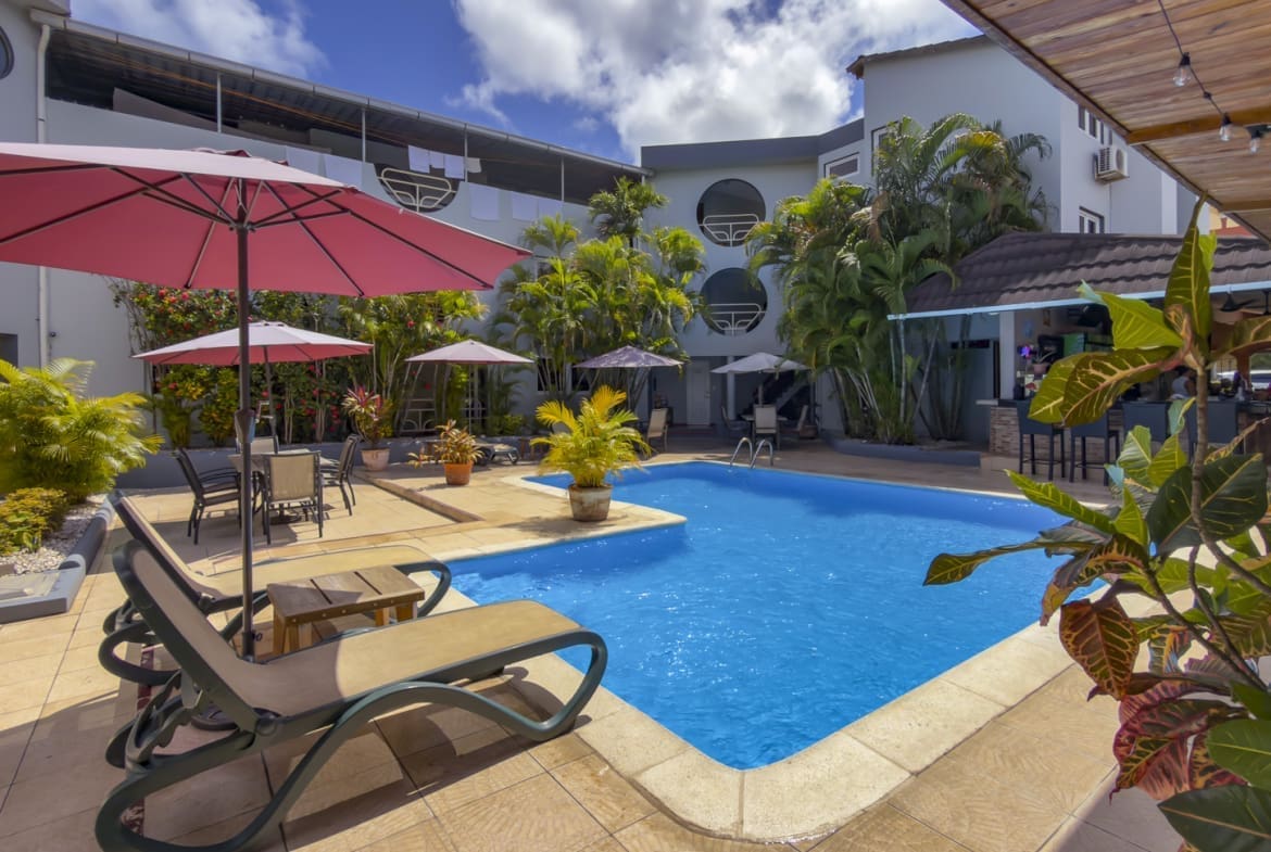 Sunny hotel pool area with pink umbrellas, lounge chairs, and tropical plants around a blue rectangular pool.
