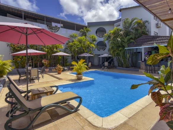 Sunny hotel pool area with pink umbrellas, lounge chairs, and tropical plants around a blue rectangular pool.