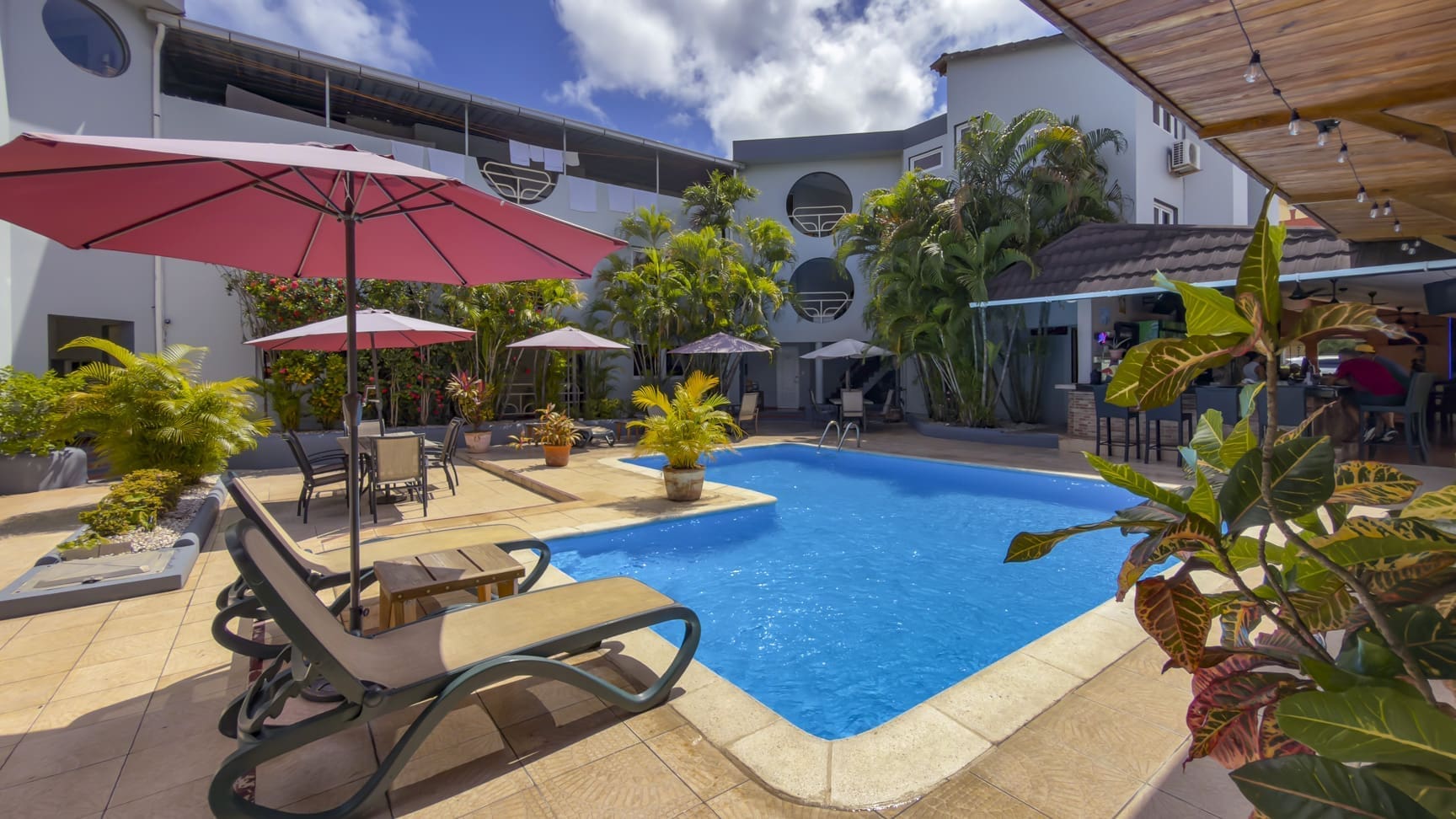 Sunny hotel pool area with pink umbrellas, lounge chairs, and tropical plants around a blue rectangular pool.